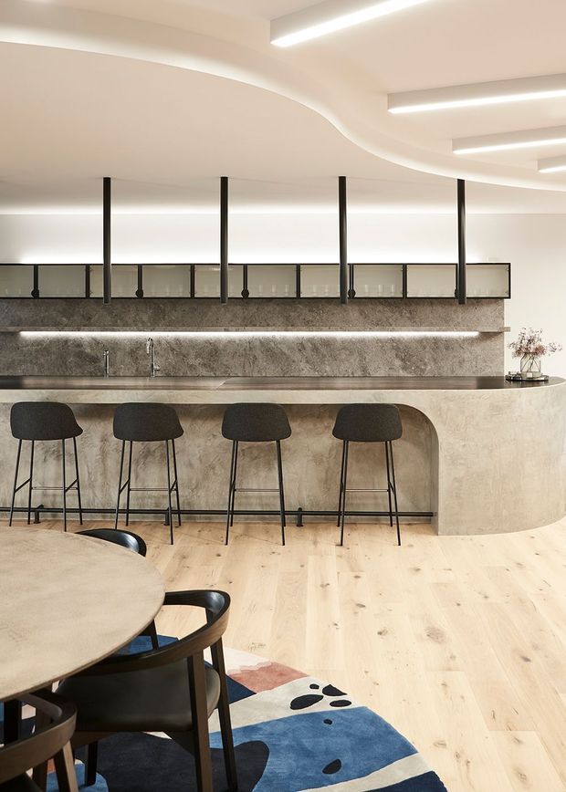 A refined open-plan kitchen and dining area with a long curved island and integrated stone-look benchtop, four dark upholstered bar stools, pale timber flooring, and a partially visible round dining table. The kitchen features a textured grey stone backsplash, brushed metal tapware, dark-framed glazed overhead cabinetry, concealed linear lighting, and a sculptural undulating white ceiling with recessed strip lights. A colourful abstract rug adds a small accent beneath the dining area.