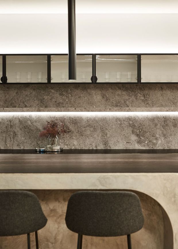 Close-up of a refined kitchen or bar counter with a dark stone-look benchtop, pale veined stone backsplash and peninsula, integrated linear lighting, dark framed reeded-glass overhead cabinetry, and two upholstered stools. A slim dark cylindrical pendant hangs centrally, creating a warm, sophisticated composition.