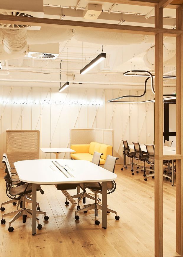 A bright, contemporary open-plan office interior viewed through a timber-framed glazed partition. The space features light timber flooring, white geometric wall panelling, exposed white services and ductwork, linear and sculptural pendant lighting, a large white meeting table with ergonomic office chairs, smaller worktables, a yellow sofa, and acoustic screening elements.