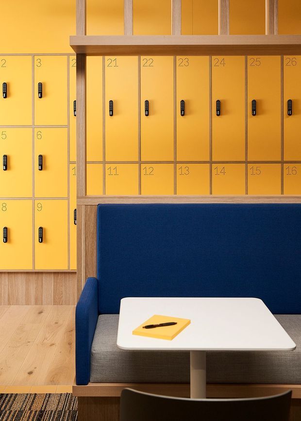 A warmly lit contemporary interior featuring a built-in yellow locker wall with black handles and numbered doors, framed by light timber slatted joinery. In the foreground is a navy-blue upholstered booth with a white table, grey seat cushion, and dark timber chair, set over light timber flooring and a patterned rug.