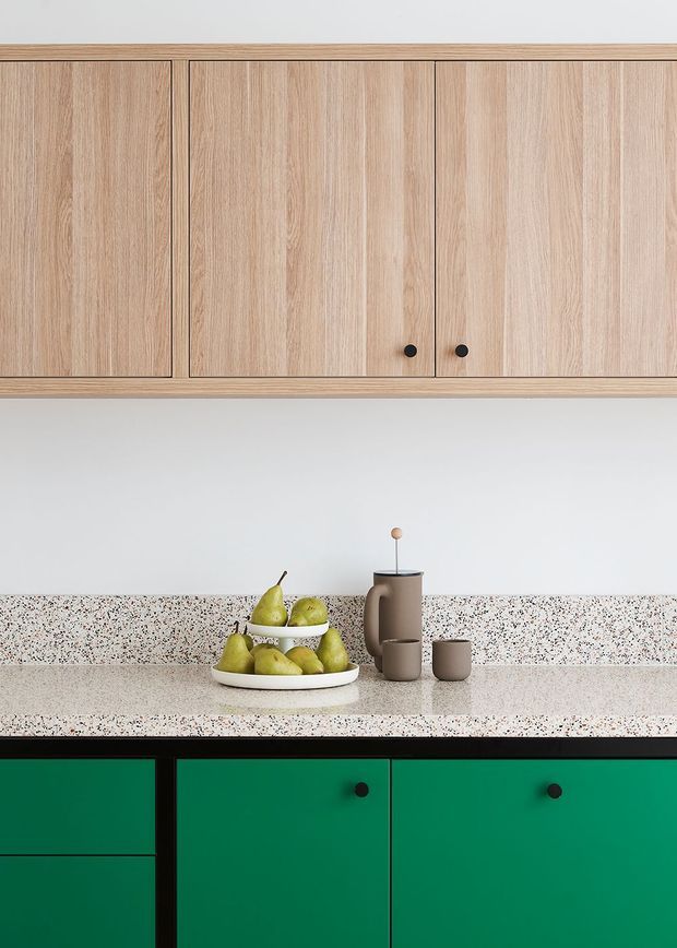 Close-up view of a contemporary kitchen featuring natural light-wood overhead cabinetry, a pale terrazzo benchtop and matching splashback, and bold deep-green lower cabinetry with black hardware. A fruit bowl and neutral-toned kitchenware sit on the counter.