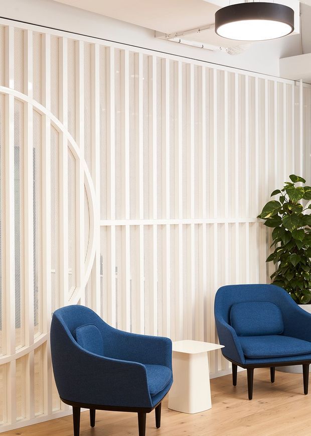 Bright contemporary interior seating area with two deep blue upholstered lounge chairs around a small white side table. The space features pale timber flooring, white vertical slatted and perforated wall screening with a large curved decorative frame, exposed ceiling services, and a large black circular pendant light. A leafy indoor plant adds greenery at the edge of the room.