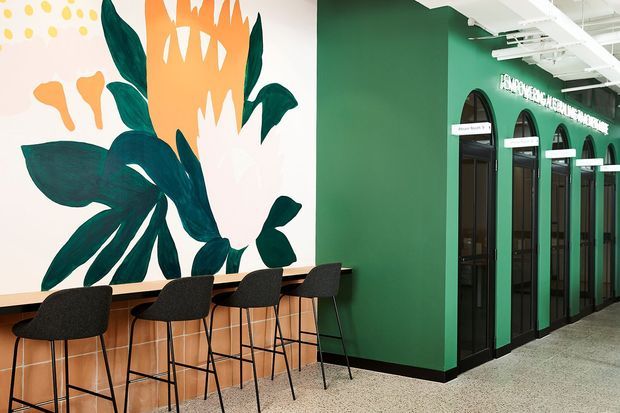 A bright contemporary commercial interior with a long built-in timber-toned counter and black bar stools beneath a large botanical mural. A deep green wall with repeated arched black-framed doorways and white signage runs along the right side. The exposed white ceiling services and polished speckled floor reinforce a modern hospitality or workplace setting.