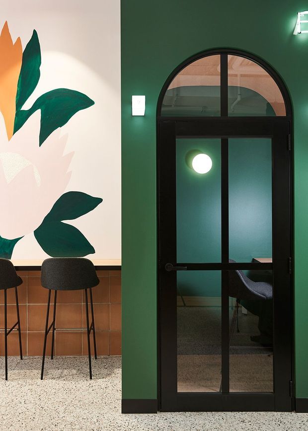 Interior view of a compact hospitality or commercial space featuring a deep green wall, black-framed arched glazed doorway, terrazzo flooring, and a large botanical mural beside a built-in counter with dark bar stools. The doorway reveals a small teal-toned room beyond, with a round wall light and seating visible through the glass.