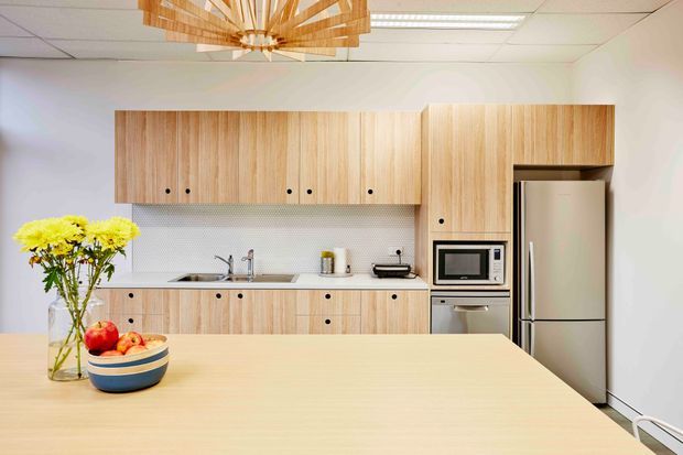 Bright contemporary kitchen with pale timber cabinetry, integrated stainless-steel appliances, a long island or peninsula in the foreground, and a white geometric backsplash. A timber slatted pendant light adds a warm architectural feature above the workspace.