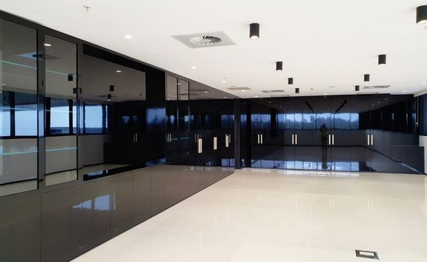 A large, polished commercial interior with extensive glossy black built-in cabinetry and glass-fronted partitions along the walls. The open floor is finished in a pale reflective surface, while a white ceiling contains recessed air-conditioning grilles and evenly spaced black cylindrical pendant downlights. The space appears clean, minimal, and highly reflective, with windows or glazed partitions along the far side.