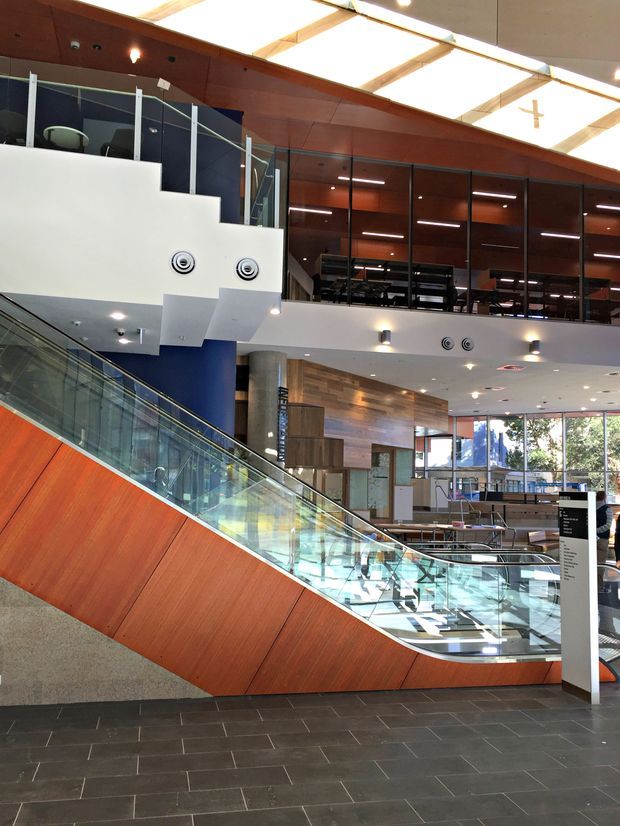 A spacious multi-level contemporary commercial interior with a prominent glass-sided escalator, warm timber wall and ceiling finishes, dark tiled flooring, extensive internal glazing, exposed services, and a large skylit atrium. Seating, counters, and an information directory are visible in the lower-level public area.