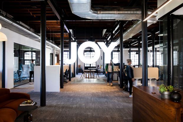A bright, open-plan modern office interior with exposed black structural columns, visible ductwork, suspended lighting, glazed meeting rooms, carpeted flooring, workstations, and timber reception or storage joinery. Large illuminated “JOY” lettering forms a prominent feature in the central workspace.