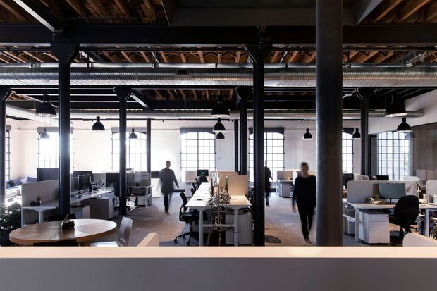 A spacious open-plan office in a converted industrial-style interior, with exposed timber roof framing, black steel columns and beams, visible ductwork, large factory-style windows, pendant lighting, and rows of contemporary workstations separated by low partitions.