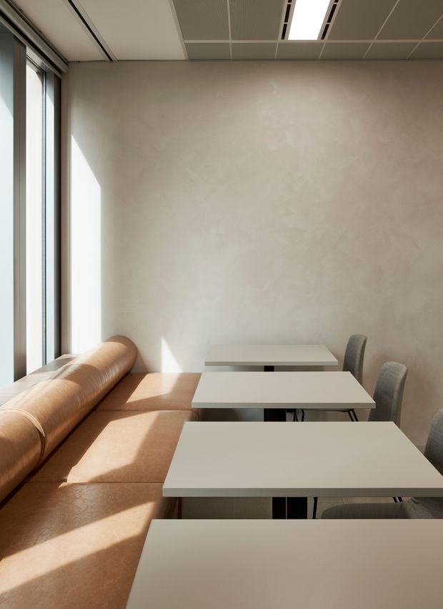 A bright, minimalist commercial dining or café interior with a long tan upholstered banquette along full-height windows, several small white rectangular tables, and simple grey dining chairs. The room has a pale textured plaster wall, a suspended acoustic ceiling with recessed linear lighting, and strong warm sunlight creating geometric shadows across the seating and floor.