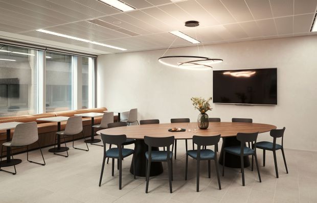Contemporary commercial breakout and meeting area with a large oval timber meeting table surrounded by black-framed chairs, additional bar-height seating along full-height glazing, a wall-mounted screen, and sculptural pendant lighting. The space has a light tiled floor, white walls, and a suspended acoustic grid ceiling.