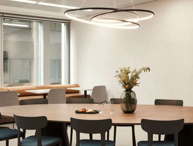 Contemporary office dining and collaboration area with a long light-wood table, dark upholstered chairs, a built-in banquette by the windows, and a sculptural looping pendant light. The space has white walls, a suspended ceiling, expansive glazing, and a restrained neutral palette with a floral arrangement as a central table accent.