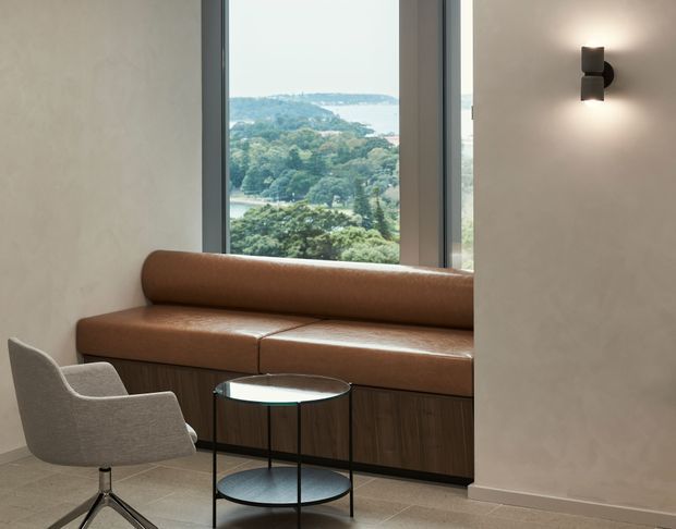 A refined contemporary sitting area with a built-in tan leather banquette beneath tall windows, paired with a light upholstered armchair and a round black-framed glass coffee table. Dark timber cabinetry forms the base of the banquette, while a minimalist wall sconce illuminates the textured light wall. The windows overlook a lush, hilly landscape and water beyond.