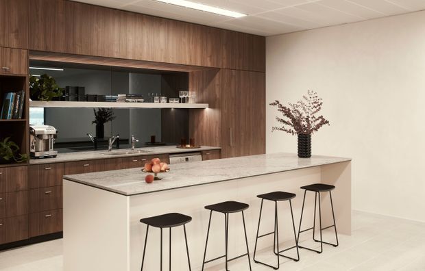 A contemporary kitchen with a large light stone island and seating for four, set against full-height dark timber cabinetry. The rear wall includes open shelving, a reflective dark splashback, integrated appliances and a sink, while simple black stools and a vase provide restrained accents. The space has a clean, neutral palette and a suspended grid ceiling with bright linear lighting.