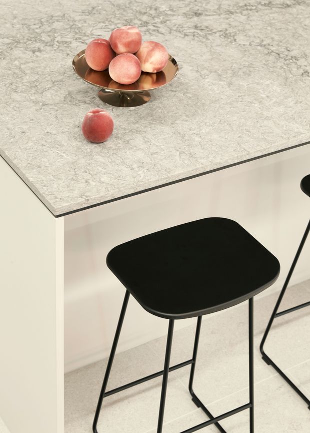 Close-up view of a contemporary kitchen island or breakfast bar with a pale grey veined stone benchtop, light neutral cabinetry, and black minimalist bar stools. A metallic fruit bowl with peaches sits on the counter, and pale tiled flooring is visible below.