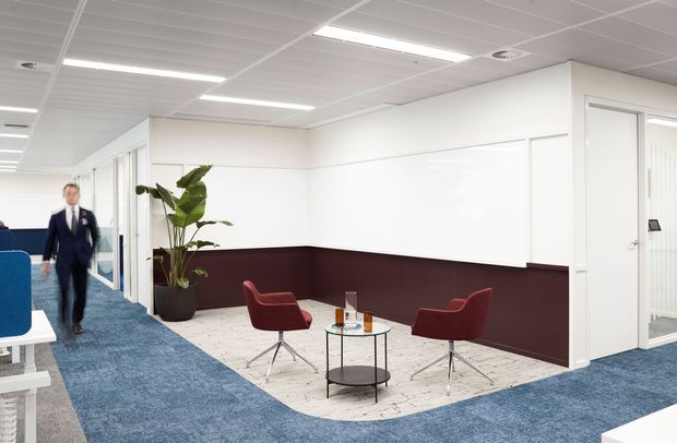 A bright contemporary commercial office interior with an open corridor, blue carpet, white walls and suspended ceiling, and a small informal meeting or waiting area. Two burgundy lounge chairs surround a round glass coffee table on a light patterned rug, with a potted plant nearby. Glazed office partitions and doors line the corridor, and a person is walking in the background.