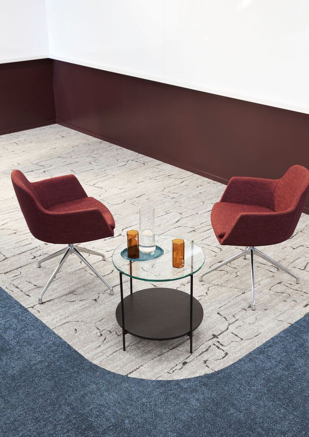 A refined contemporary lounge setting with two burgundy upholstered armchairs arranged around a small round two-tier coffee table. The room features white upper walls, a dark burgundy lower wall panel, and a large light patterned area rug with a rounded edge over blue carpet. Clear glass, black metal, and amber decorative objects sit on the table.