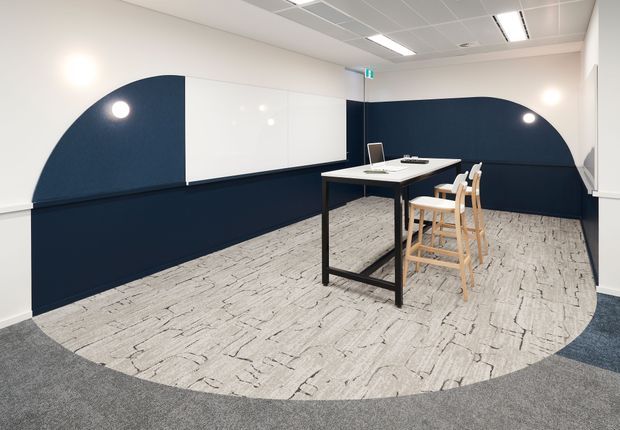 A compact contemporary commercial meeting or collaboration area with a tall black table, stools, and a laptop. The space features dark navy curved wall panels, large whiteboards, light patterned carpet with a rounded inset shape, white walls, and a suspended grid ceiling with integrated fluorescent lighting.