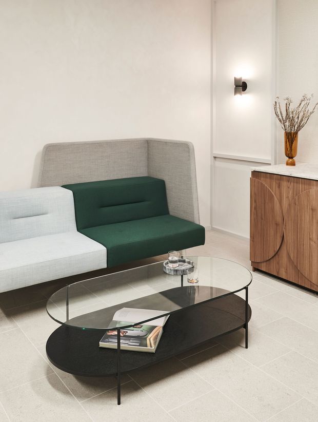 Contemporary living room with a pale upholstered sectional sofa featuring a deep green seat section, a black oval two-tier coffee table with glass top, and a curved timber sideboard with a light stone benchtop. The space has pale tiled flooring, white decorative wall panelling, and a minimalist wall sconce, creating a refined neutral setting with warm wood accents.