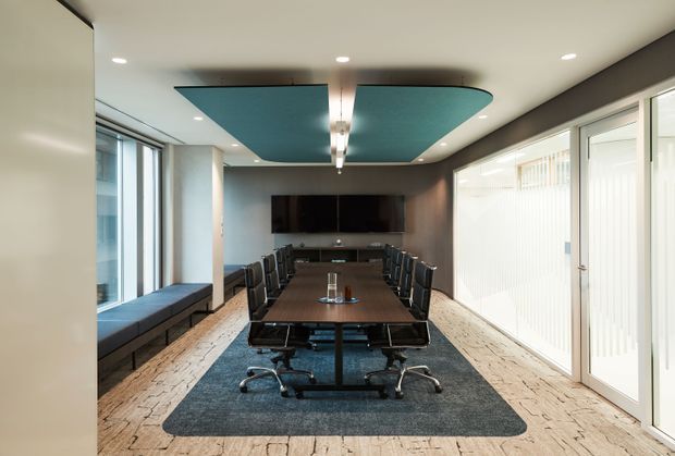 Contemporary boardroom with a long dark wood conference table, black office chairs, wall-mounted display, built-in window seating, and expansive glazed doors and windows. A distinctive teal acoustic ceiling feature and dark carpet contrast with pale textured flooring and neutral walls.