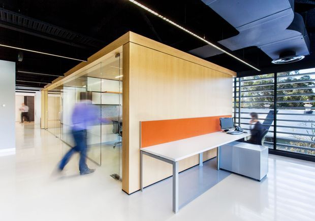 Contemporary open-plan office interior with timber-lined enclosed meeting or work pods, extensive glass partitions, white flooring, and shared workstations. A vivid orange wall panel provides contrast against the pale timber and white surfaces. The exposed dark ceiling contains linear strip lighting and visible mechanical services, while large louvred windows bring in daylight and overlook greenery.