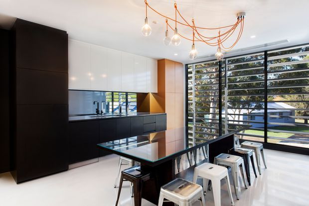 Contemporary open-plan kitchen with a large black island and bar seating, glossy white overhead cabinets, dark integrated cabinetry, and a reflective dark splashback. Floor-to-ceiling louvred glazing opens views to the landscaped exterior. Exposed orange electrical cords suspend multiple pendant bulbs, creating a playful industrial accent in an otherwise sleek, high-contrast space.