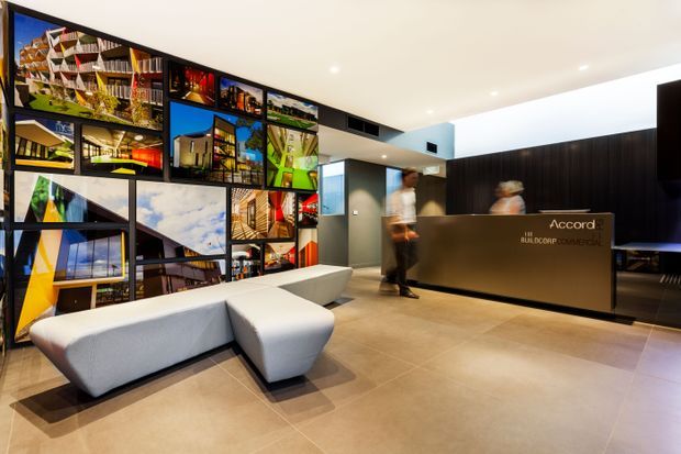 Contemporary hotel or commercial reception lobby with a large illuminated reception desk, dark timber wall backdrop, tiled flooring, sculptural pale lounge seating, and a dramatic floor-to-ceiling collage of architectural photographs. Staff members are visible behind and beside the desk.
