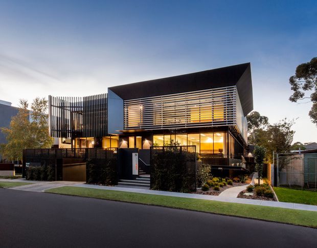 A contemporary multi-storey residential facade photographed at dusk. The house has a strongly geometric form with a dramatic projecting dark upper roof or canopy, extensive glazing, horizontal external sun-shading slats, and a vertical screen along one side. Dark fencing, a front entry stair, landscaped planting, and a curved path sit between the building and the street.
