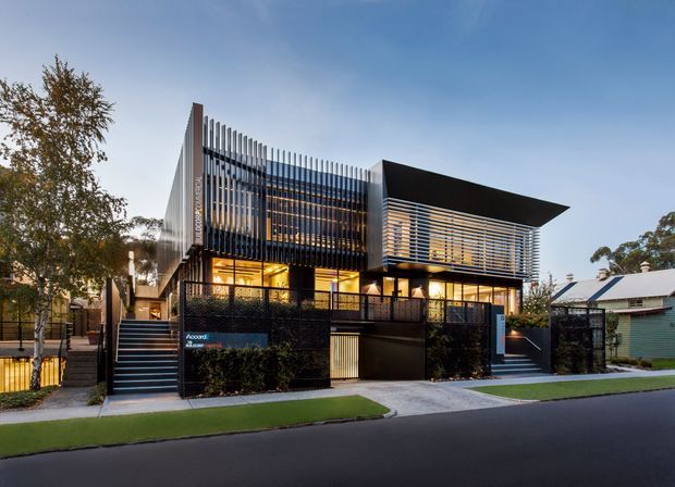 Street-level exterior view of a contemporary multi-storey building with a strongly articulated dark facade, vertical metal fins, horizontal sun-shading screens, extensive glazing, and a projecting flat roof canopy. Landscaped frontage, stairs, fencing, and neighbouring residential buildings are visible.