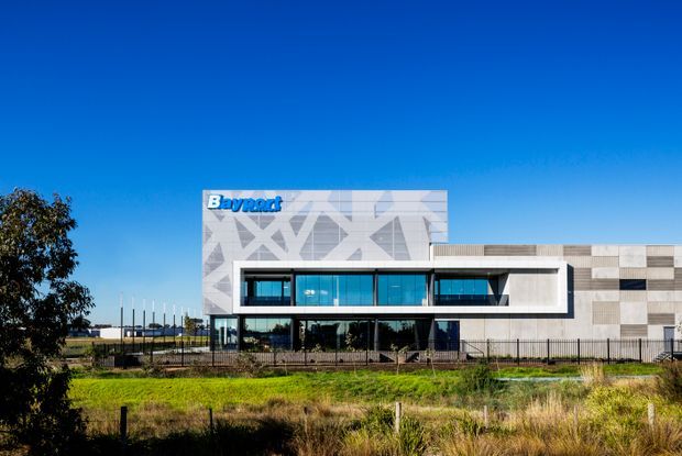 Exterior photograph of a contemporary Bayrad commercial or institutional building under a clear blue sky. The facade combines a prominent white geometric-patterned upper volume, extensive dark-framed glazing, and a long wall clad in varied pale grey and beige rectangular panels. The building sits behind fencing with landscaped grass in the foreground.