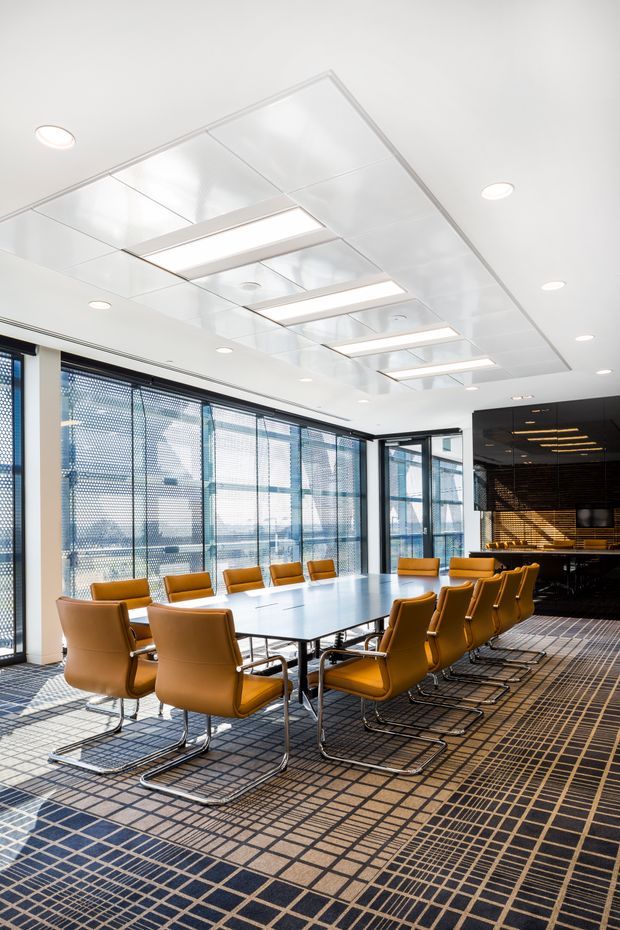 Bright contemporary commercial meeting room with a large rectangular conference table surrounded by tan leather cantilever chairs. The space has extensive floor-to-ceiling glazing with patterned blinds, a highly reflective white suspended ceiling with recessed linear lighting, and a dark built-in bar or kitchenette at the rear. A dark geometric carpet covers the floor.