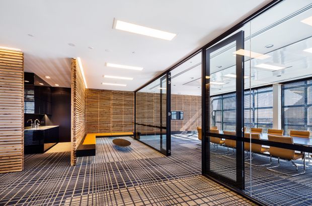 Contemporary commercial office interior with a glazed meeting room, open-plan workspace, compact kitchenette, timber slatted screening, black-framed glass partitions, and patterned carpet tiles. The space is bright, highly structured, and defined by strong linear grid patterns and warm timber accents.