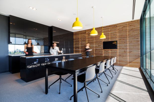 A contemporary open-plan commercial breakout and dining area with a long black communal table, white chairs, dark glossy kitchen or service cabinetry, and a timber slatted feature wall. Bright yellow pendant lights provide a strong accent, while full-height glazing lines the right side and daylight fills the space. People are present but the focus is the interior fit-out.