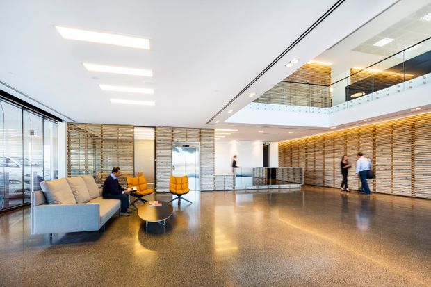 Bright contemporary commercial lobby or reception area with a polished terrazzo-like floor, large glazed perimeter, extensive horizontal timber slat wall screens, a reception desk, lounge seating, and a mezzanine or upper-level opening with glass balustrading. Linear ceiling lighting and integrated warm wood detailing create a refined, spacious workplace entrance.