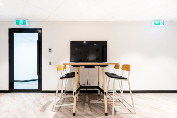 Bright, minimalist commercial interior featuring a high communal table with stools arranged around a wall-mounted television. The room has white walls, a suspended white ceiling, pale timber-look flooring, black skirting and door framing, and an emergency exit door.