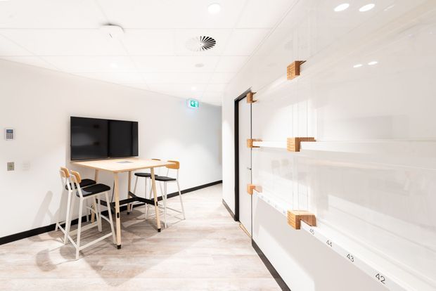 Bright, minimalist commercial interior resembling a showroom, educational space, or collaborative workspace. The room has white walls and ceiling, pale timber-look flooring, a wall-mounted black display screen, a high light-wood table with white stools, and extensive white display shelving with small timber brackets. Recessed lighting, exposed ceiling services, black skirting, and a glass door contribute to a clean contemporary fit-out.