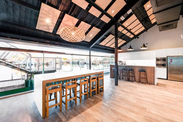 An open-plan commercial-style bar and dining area with a long timber communal table and matching stools, a white bar counter with timber cabinetry, stainless-steel appliances, and extensive glazing overlooking a waterfront or marina-like setting. The space features an exposed dark ceiling with black steel framing, acoustic or illuminated ceiling panels, pendant lighting, and polished timber-look flooring.