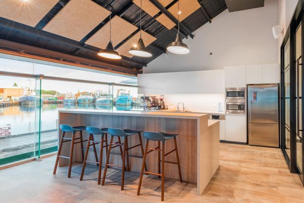 A contemporary open-plan kitchen and bar overlooking a marina or waterfront through extensive glazing. The space features a large island with a timber-look base and pale benchtop, four stools, integrated stainless-steel appliances, white vertical-panel cabinetry, timber-look flooring, black exposed ceiling structure, acoustic ceiling panels, and suspended pendant lights. Full-height glass and black-framed glazing create a bright connection to the waterfront.