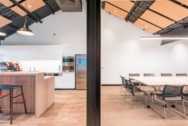 A bright open-plan commercial interior showing a compact kitchenette and island on the left, connected to a meeting or dining area on the right. The space has white walls, pale timber-look flooring, white cabinetry, a timber-toned island, a long communal table with office chairs, exposed dark ceiling structure, suspended acoustic ceiling panels, and contemporary pendant and linear lighting.