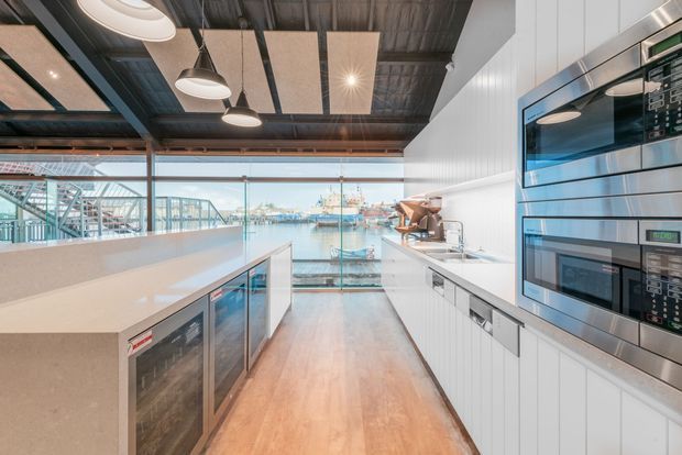 A bright contemporary commercial kitchen or hospitality fit-out overlooking a working waterfront. The space features a long pale stone island with integrated under-counter refrigeration, white vertically panelled cabinetry, stainless-steel wall ovens and appliances, warm timber flooring, extensive floor-to-ceiling glazing, and an exposed dark industrial ceiling with suspended acoustic panels and pendant lighting. Harbour infrastructure and water are visible beyond the glazing.