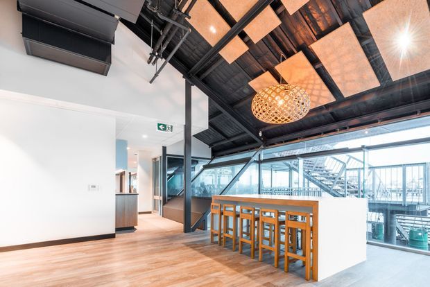 A bright contemporary commercial interior with a high exposed industrial ceiling, black structural framing, visible services, warm timber flooring, and a long timber communal bar table with stools. Full-height glazing overlooks an external stair and terrace structure. Suspended acoustic panels and a woven pendant light add warmth to the otherwise industrial fit-out.