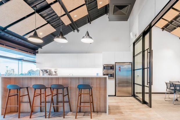 Bright contemporary commercial-style kitchen and breakout space with a long timber-look island, four bar stools, white full-height cabinetry, integrated appliances, and stainless-steel refrigerator. The space features a high exposed dark ceiling with suspended acoustic panels, black-framed glazed partitions, pendant lighting, and pale timber flooring. A seating area is visible beyond the glass partition, with expansive glazing at the left.