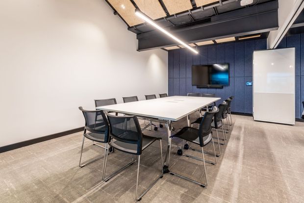Modern commercial meeting room with a large white conference table surrounded by black and grey chairs. The space features a dark exposed structural ceiling with suspended acoustic panels and linear lighting, a blue acoustic-panel feature wall with wall-mounted display, white walls, a large whiteboard, and a neutral carpeted floor.