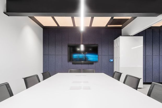 A contemporary meeting room or boardroom with a long white conference table surrounded by black mesh chairs. The rear wall is clad in dark blue-grey acoustic panels and includes a wall-mounted display, while a dark timber slatted ceiling feature with integrated lighting adds visual interest. White walls, a white partition or cabinet, and a clean minimalist fit-out create a professional commercial interior.