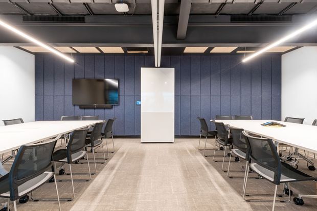 A modern commercial meeting or conference room with two long white tables arranged along the sides, surrounded by black mesh office chairs. The room features a dark blue acoustic-panel feature wall, wall-mounted display, central white built-in cabinet, exposed industrial ceiling services, linear LED lighting, and a neutral carpeted floor.