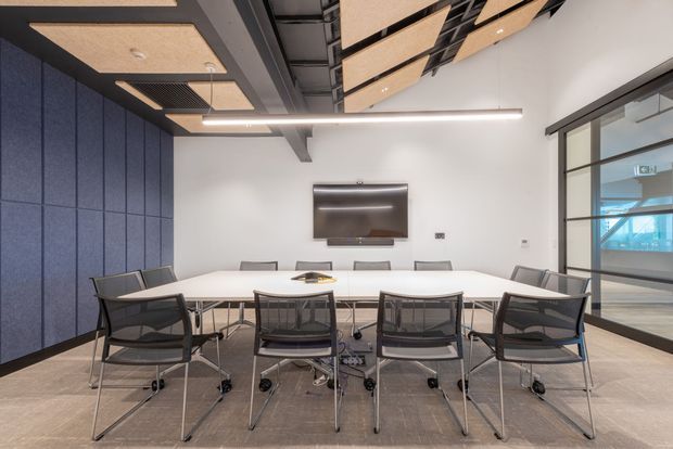 A modern commercial meeting or conference room with a long white conference table surrounded by black mesh office chairs. The room has dark blue acoustic wall panelling, a suspended acoustic ceiling with exposed services, linear LED lighting, a wall-mounted display, and a black-framed glazed partition revealing an adjacent office or circulation area. The design is clean, functional, and contemporary with a cool neutral palette.