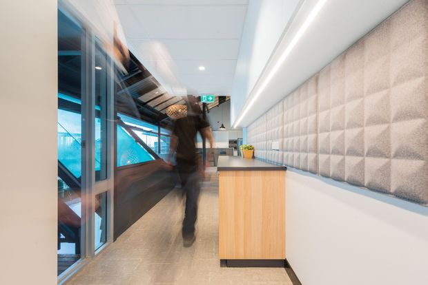 Bright contemporary commercial interior, likely an office or workplace corridor, with a long textured acoustic feature wall, integrated linear ceiling lighting, glass partitions, timber reception or storage joinery, and polished concrete-look flooring. A blurred person is walking through the space, with illuminated glazing and a visible exit sign in the background.