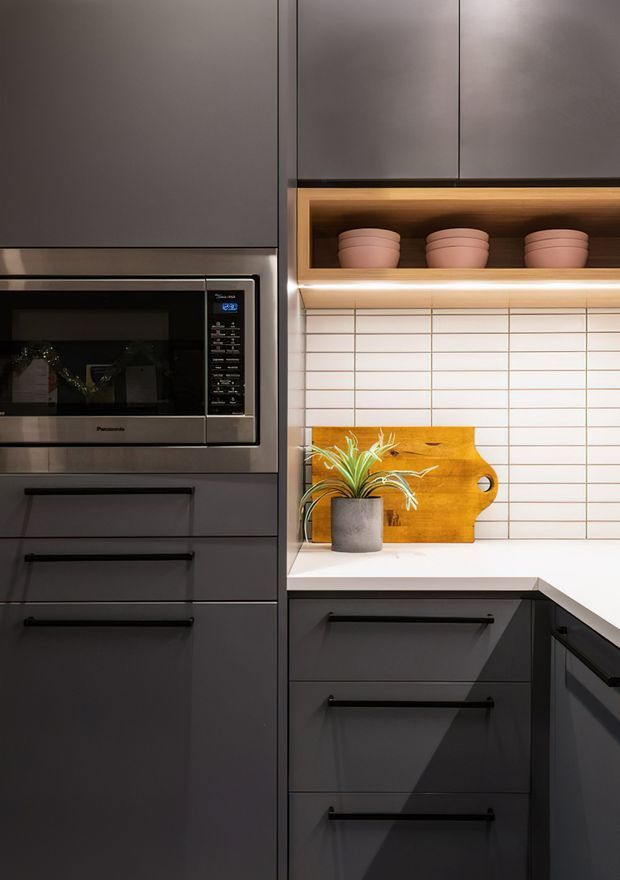 Close-up view of a contemporary kitchen with matte dark cabinetry, integrated microwave, pale benchtops, and a white horizontally gridded tiled splashback. An illuminated open shelf displays ceramic pots, while a timber cutting board and potted plant add warmth.
