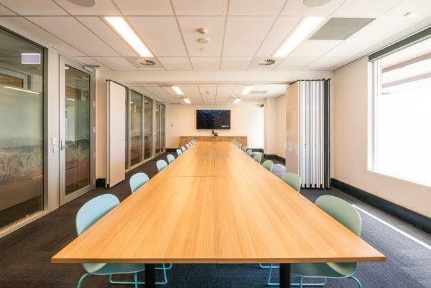 Bright contemporary commercial boardroom with a long light-wood conference table, colourful upholstered chairs, glazed meeting rooms along one side, a wall-mounted display, carpeted flooring, and a suspended acoustic tile ceiling with linear lighting.