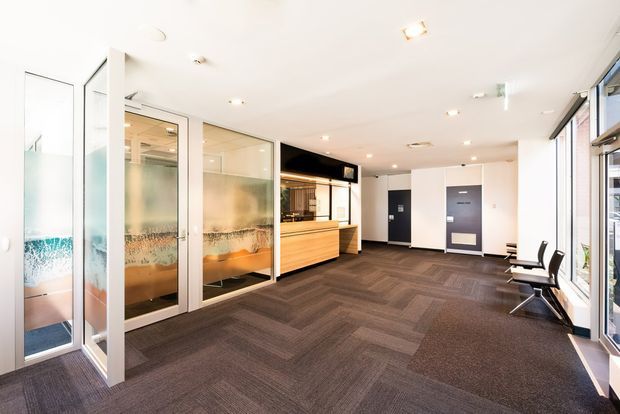 Bright contemporary commercial reception and waiting area with a timber reception desk, frosted glass meeting-room partitions, dark patterned carpet, white walls and ceiling, and a row of black seating beside full-height windows.